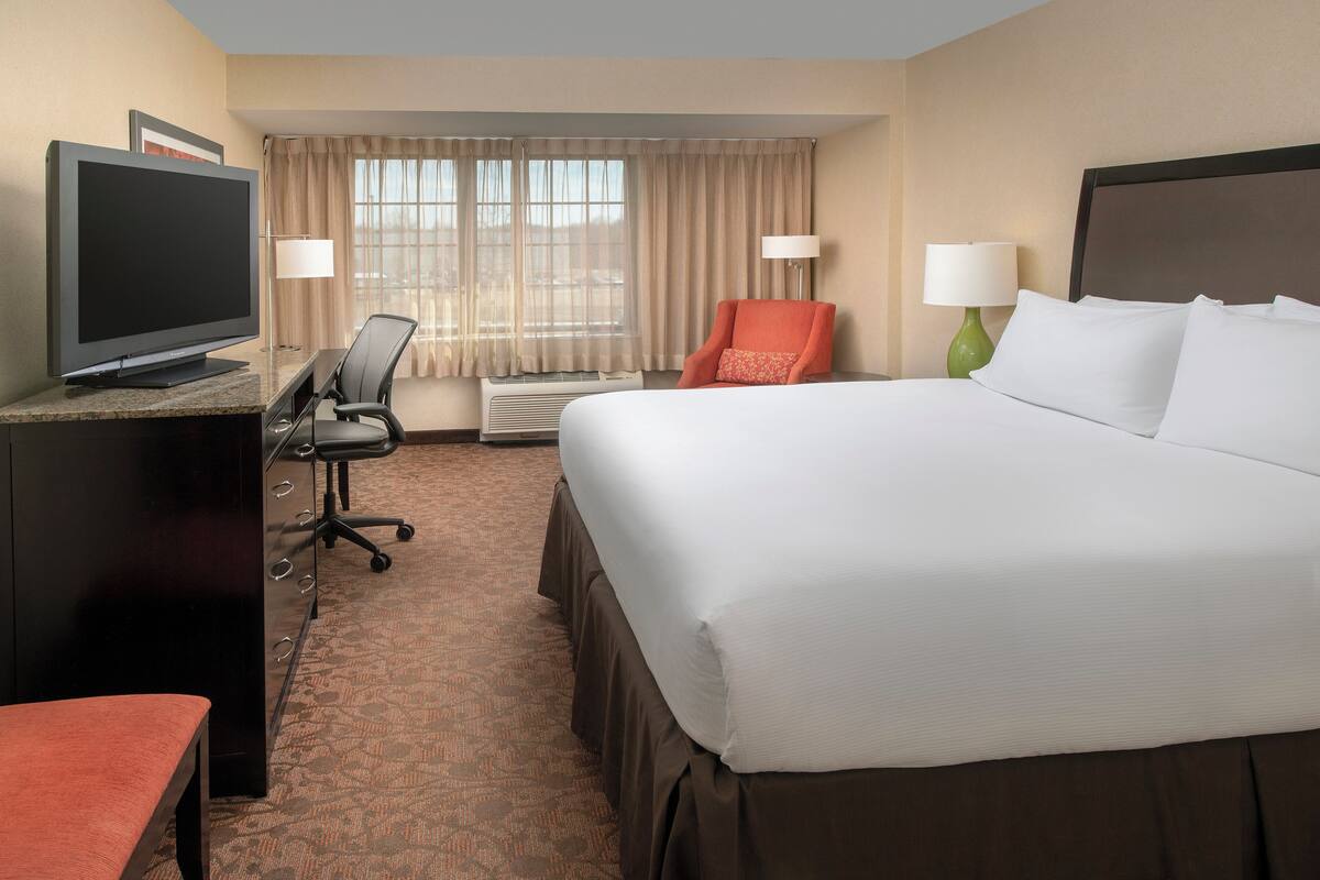 Une chambre d’hôtel avec un grand lit, un bureau doté d’un téléviseur à écran plat, une chaise, une lampe et une fenêtre avec des rideaux.