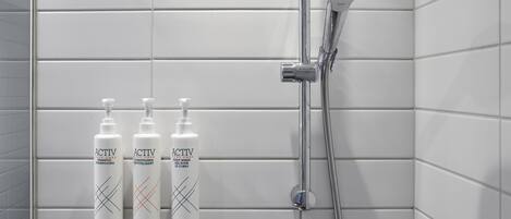Un baño con paredes de azulejos blancos, un cabezal de ducha y tres botellas de lavado corporal y acondicionador ACTIV.