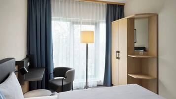 A hotel room with a bed, a desk, a chair, a lamp, and a wardrobe.