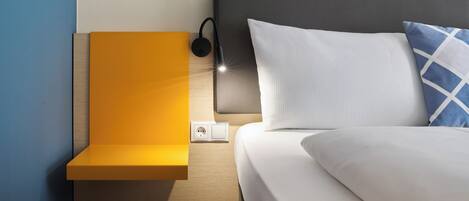 Una cama bien hecha con ropa de cama blanca, cabecera gris y almohada azul y blanca. Hay una mesita de noche pequeña con superficie amarilla y una lámpara negra.