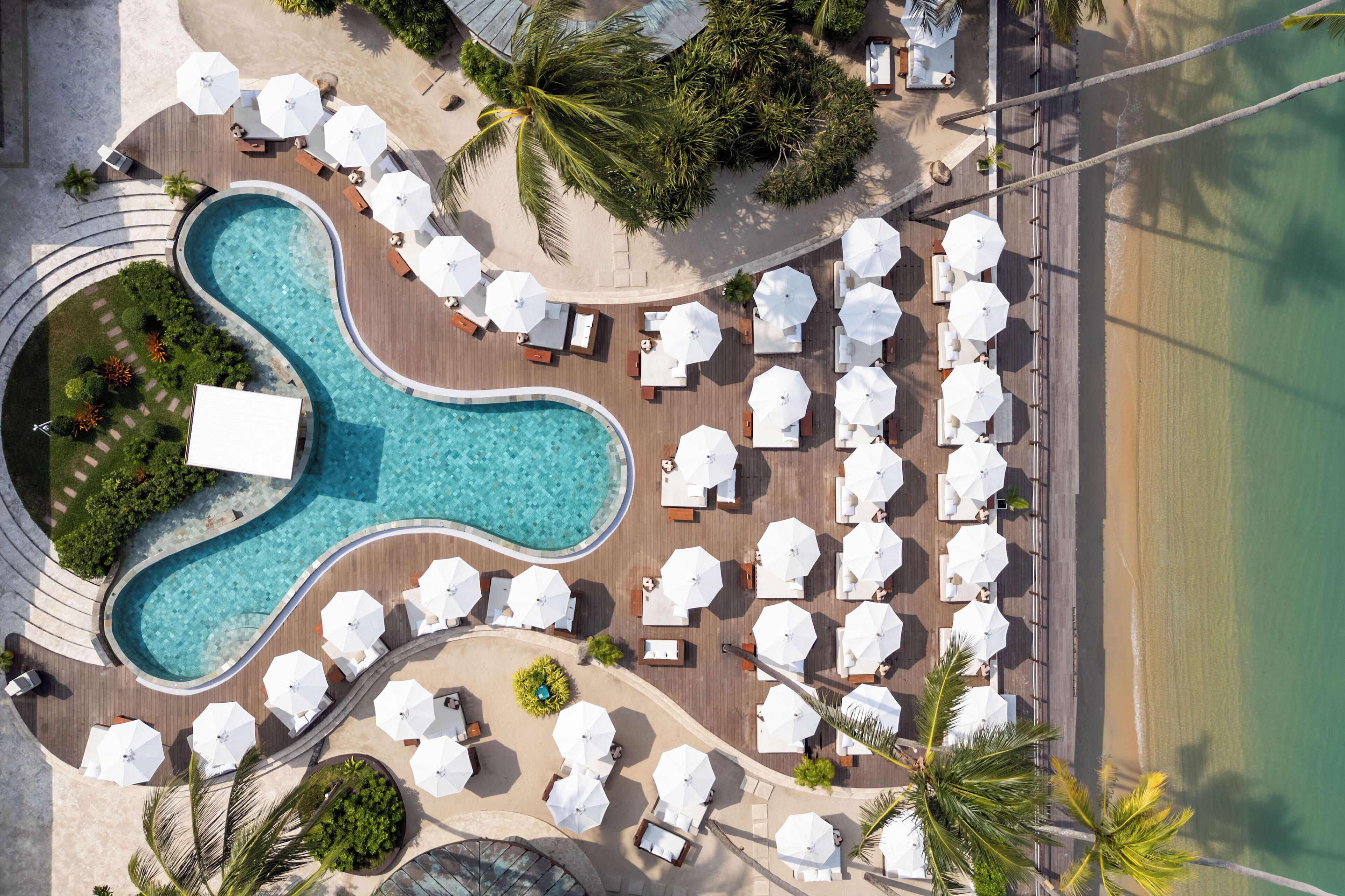 Vista aérea de un resort con piscina, tumbonas, sombrillas y una zona de playa.