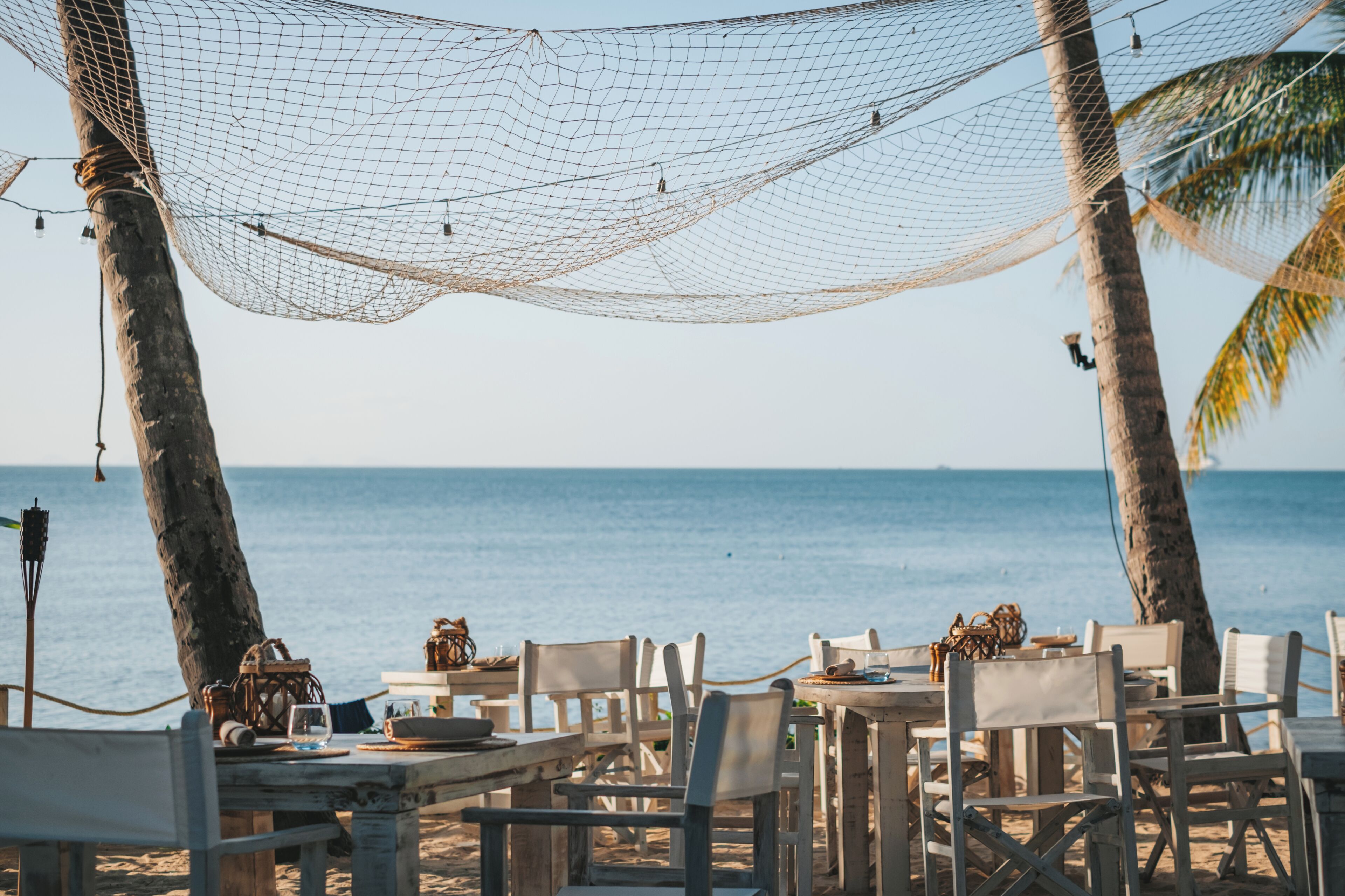 A beachside dining area with tables and chairs, a hammock, and palm trees.