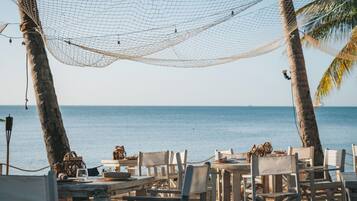 A beachside dining area with tables and chairs, a hammock, and palm trees.