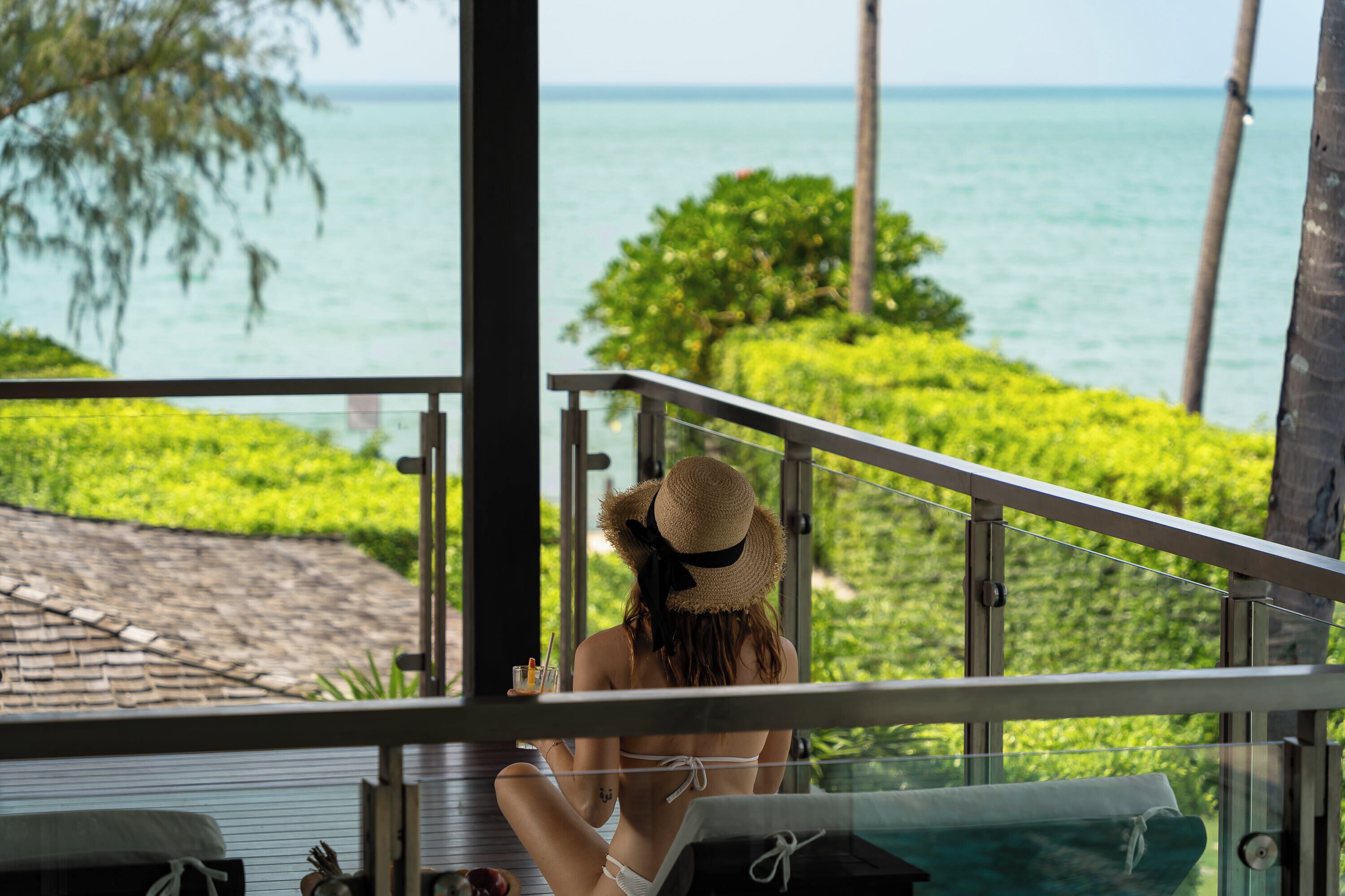 A woman in a white bikini and straw hat sitting on a balcony with a view of the ocean and greenery.