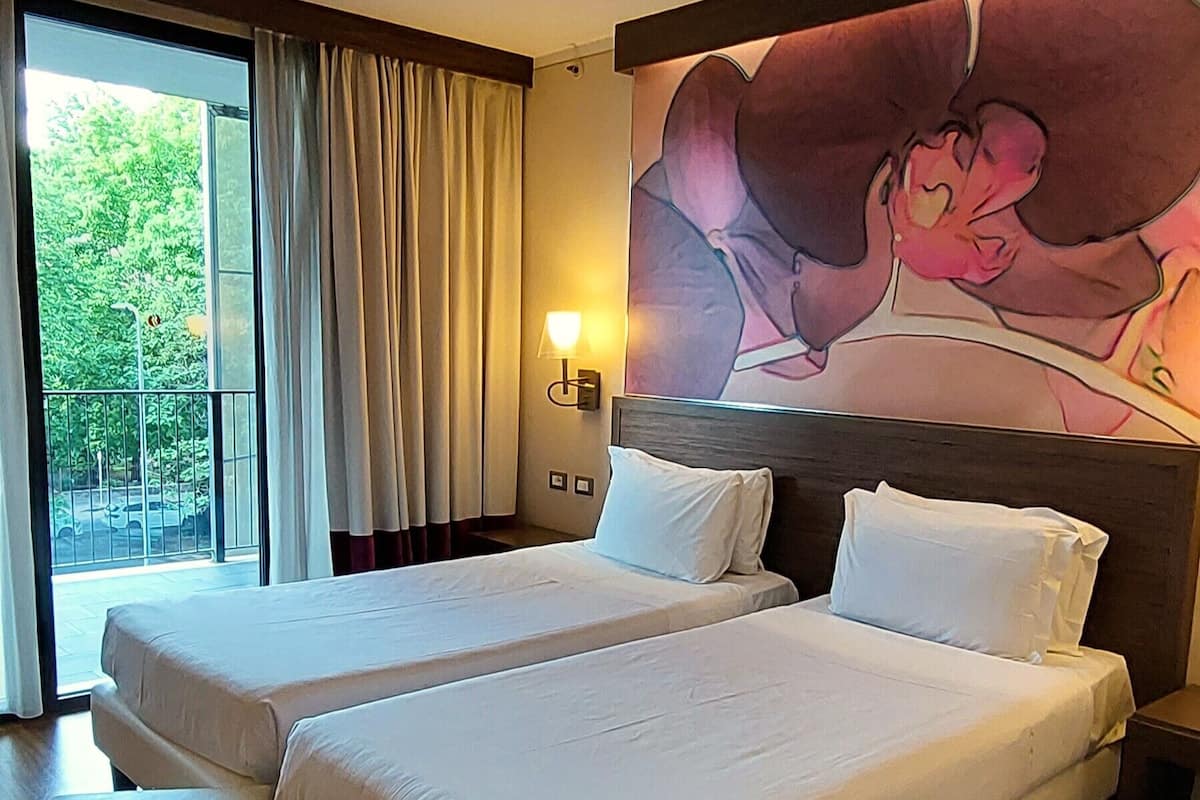 Habitación de hotel con dos camas, un ventanal con cortinas, una lámpara de pared y un cuadro de una mujer.
