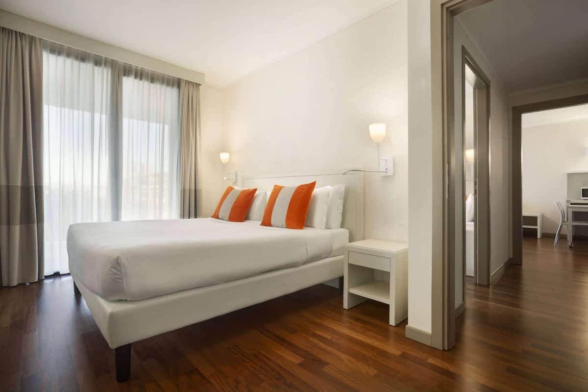 Habitación de hotel moderna con una cama grande, dos mesitas de noche y vistas a la ciudad a través de amplios ventanales.