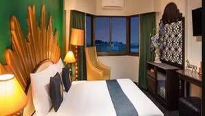 Quarto de hotel com uma cama grande, uma escrivaninha com abajur, uma cadeira e vista para o mar pela janela.