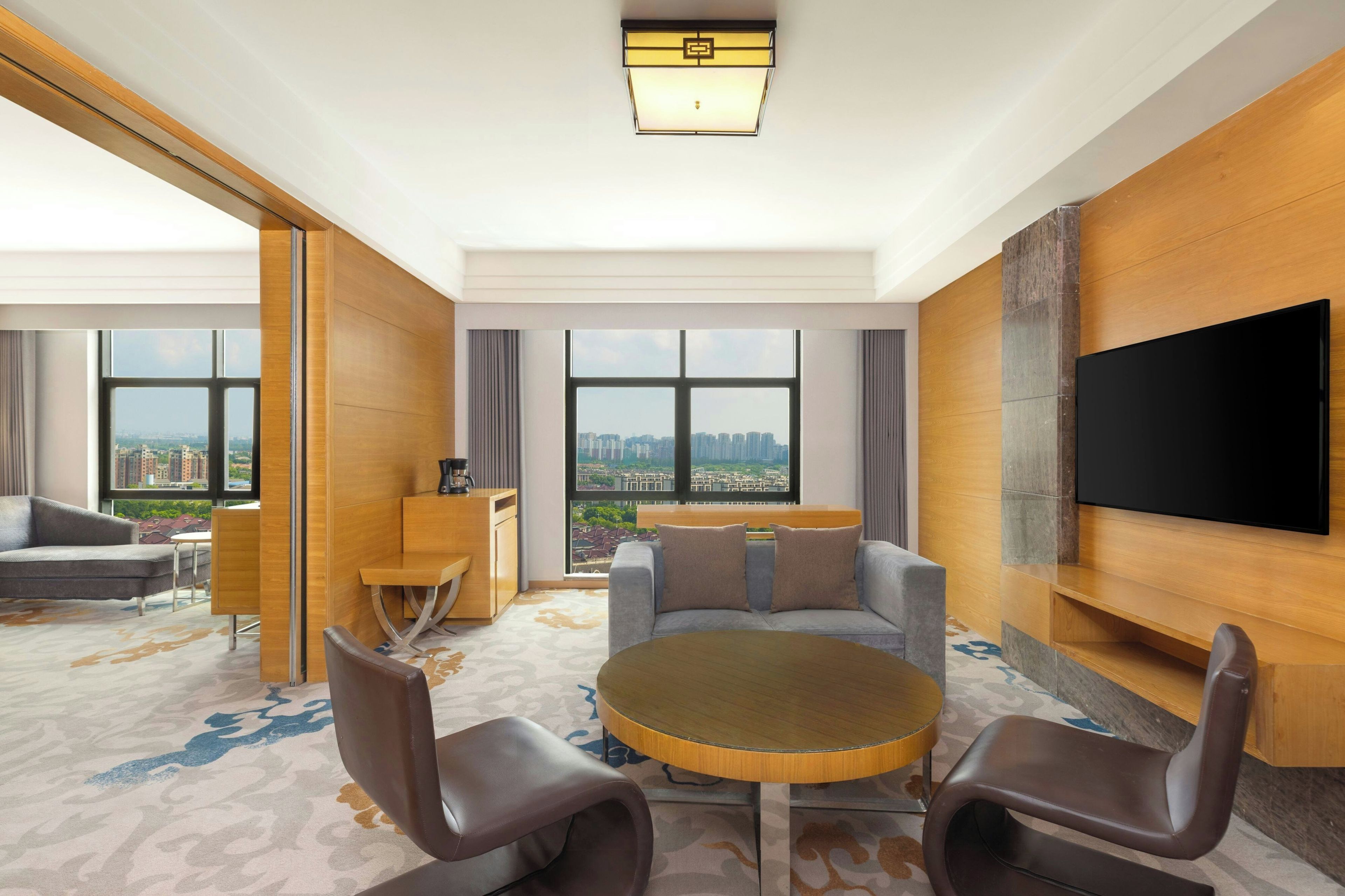 A modern hotel room with a large window, a flat-screen TV, a round table, and a sofa.