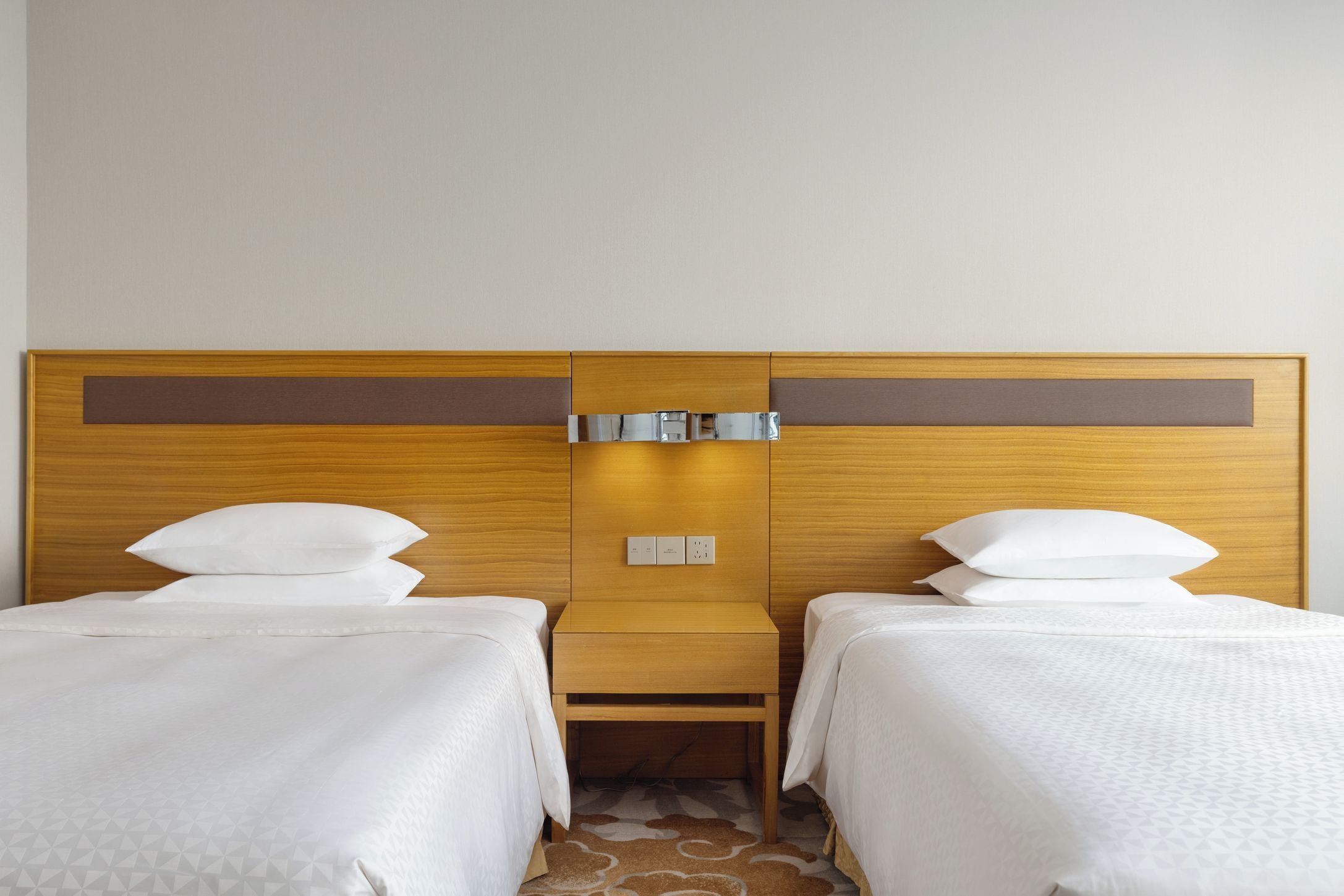 A hotel room with two single beds, a wooden headboard, a small wooden table, and a wall-mounted control panel.