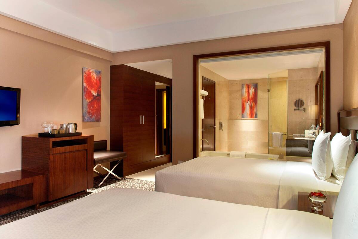 A hotel room with two beds, a desk, a TV, and a bathroom with a shower and toilet.