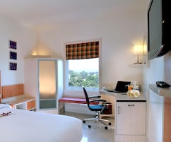 Habitación de hotel con cama, escritorio, ordenador portátil, silla, televisor y ventana con vistas a árboles.