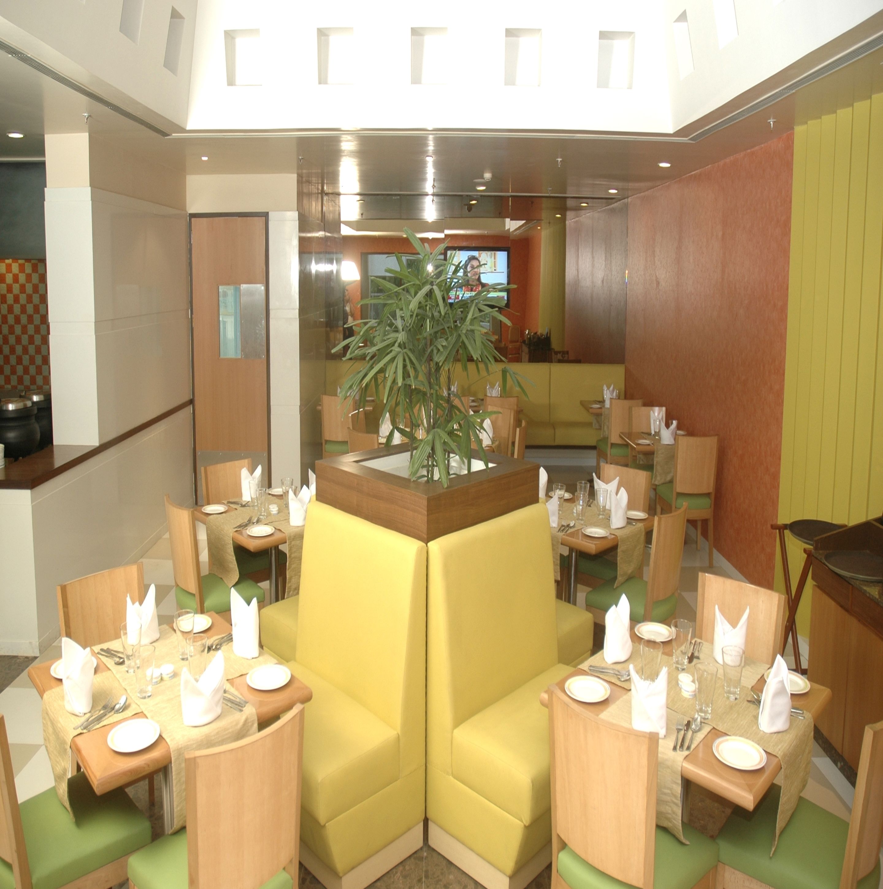 A restaurant with yellow booths, green cushioned chairs, and a central plant. Tables are set with white napkins and plates.