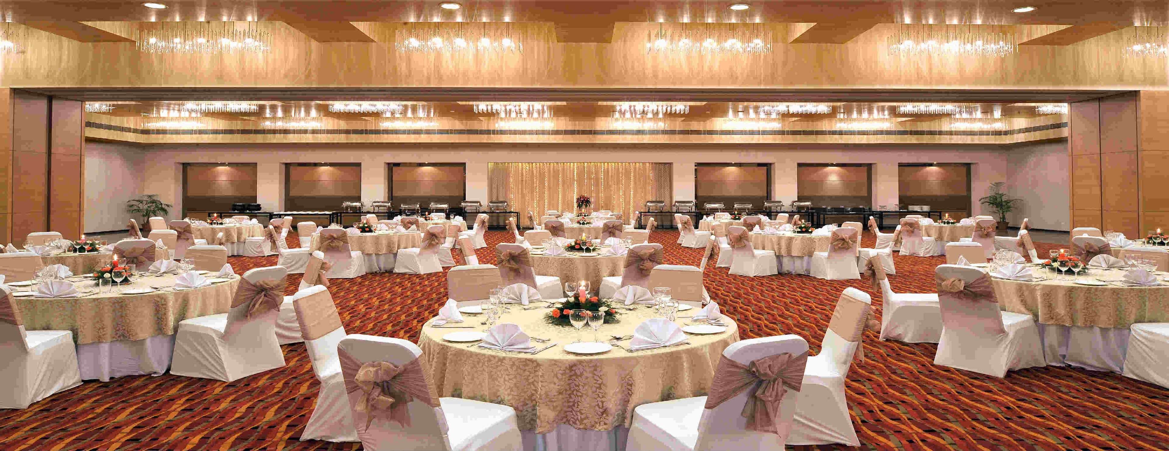 A large banquet hall with multiple round tables set for dining, each with white tablecloths and chairs, and a carpeted floor.