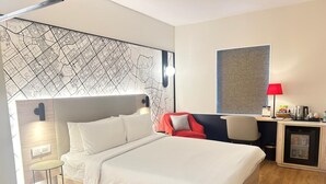 A modern hotel room with a large bed, a desk with a chair, a red armchair, a map on the wall, and a small refrigerator. - Hometel Chandigarh (Chandigarh)