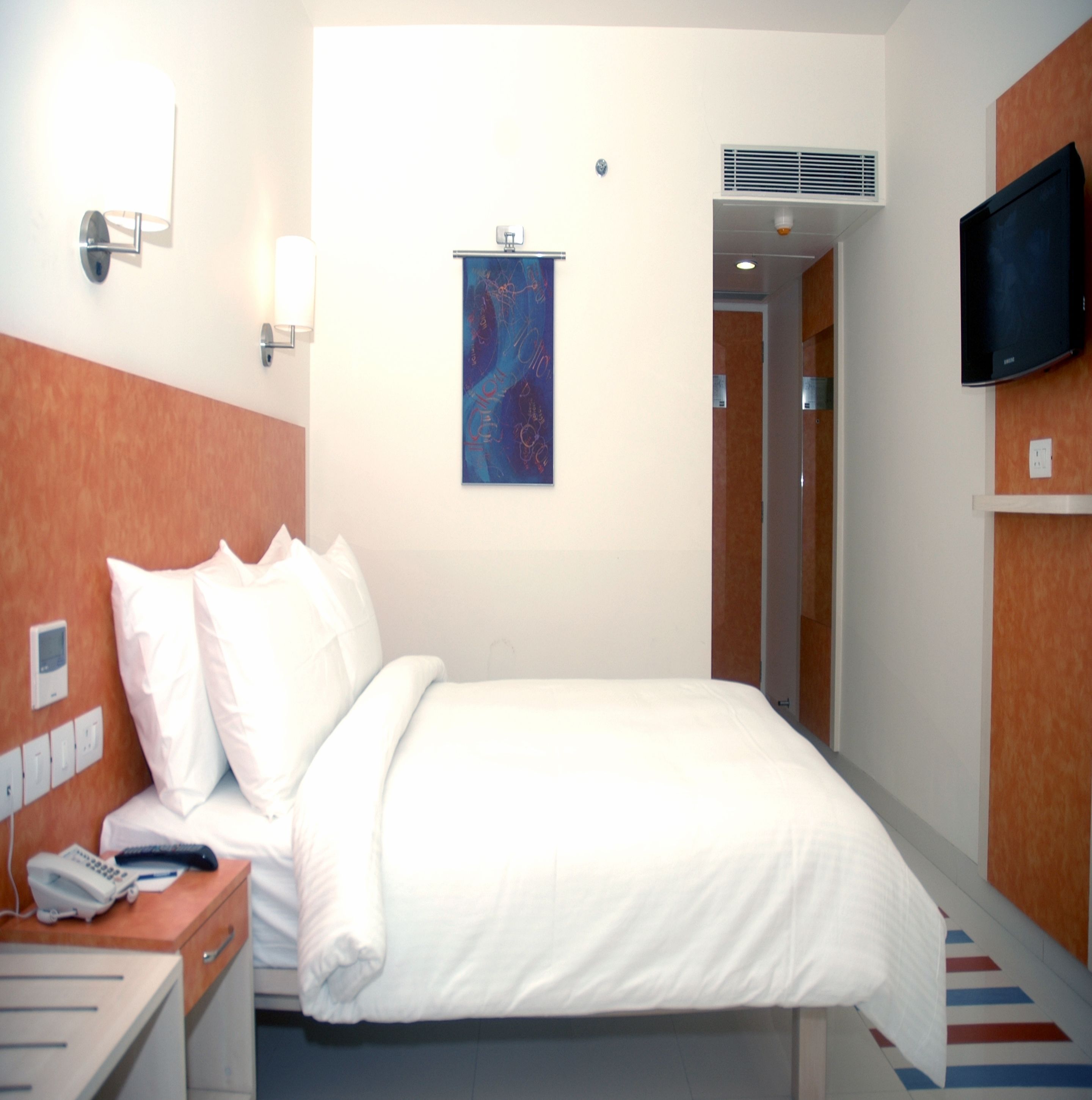 Una habitación de hotel con una cama, una mesita de noche, un televisor montado en la pared, un cuadro y una puerta que da a un baño.