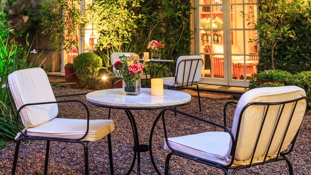 An outdoor seating area with two chairs and a table, surrounded by greenery and a building with large windows.