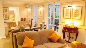 A well-lit living room with a sofa, armchairs, and a coffee table. There are framed pictures on the wall and a side table with a lamp.