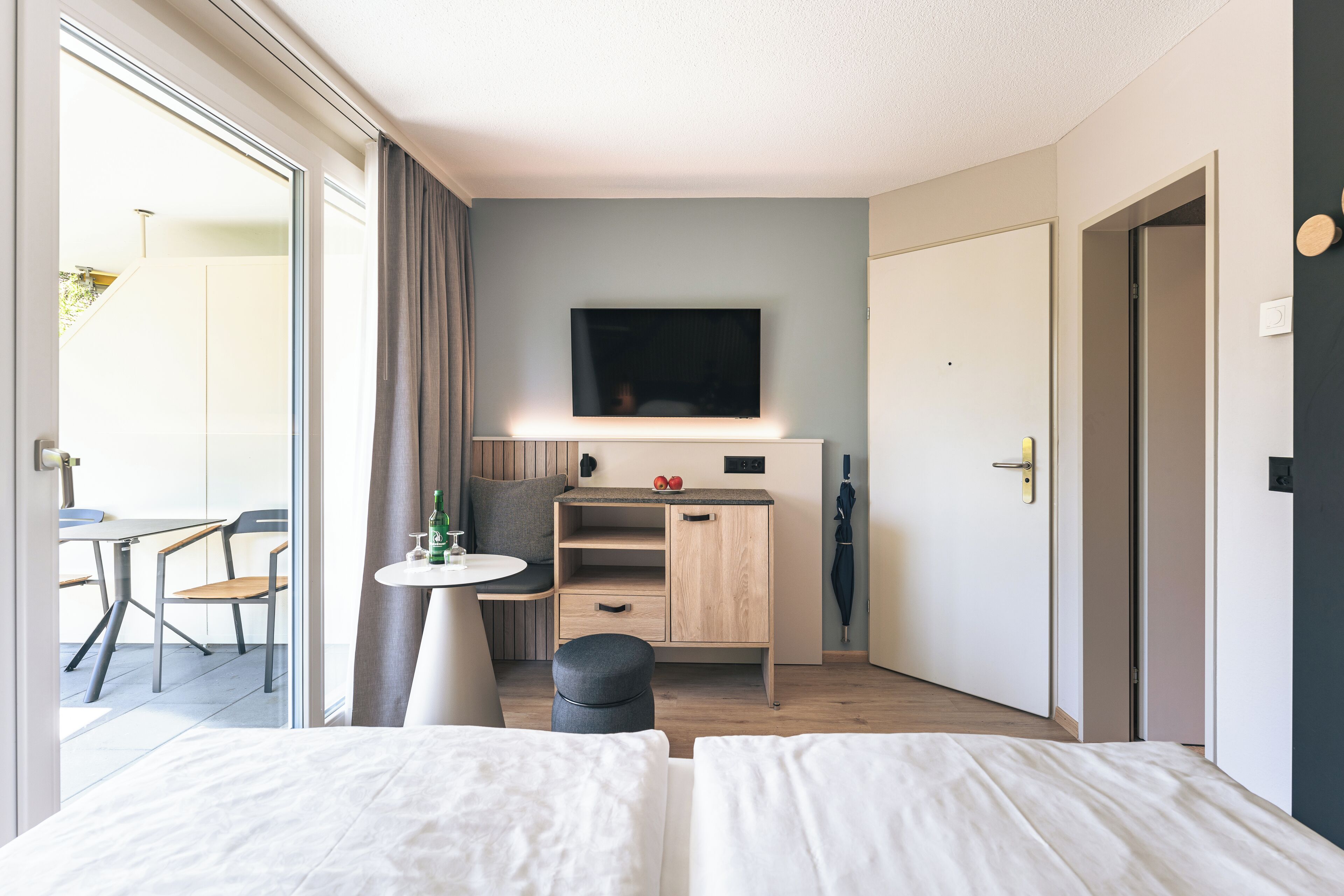 A modern hotel room with a bed, a desk with a chair, a television, and a view of the outside.