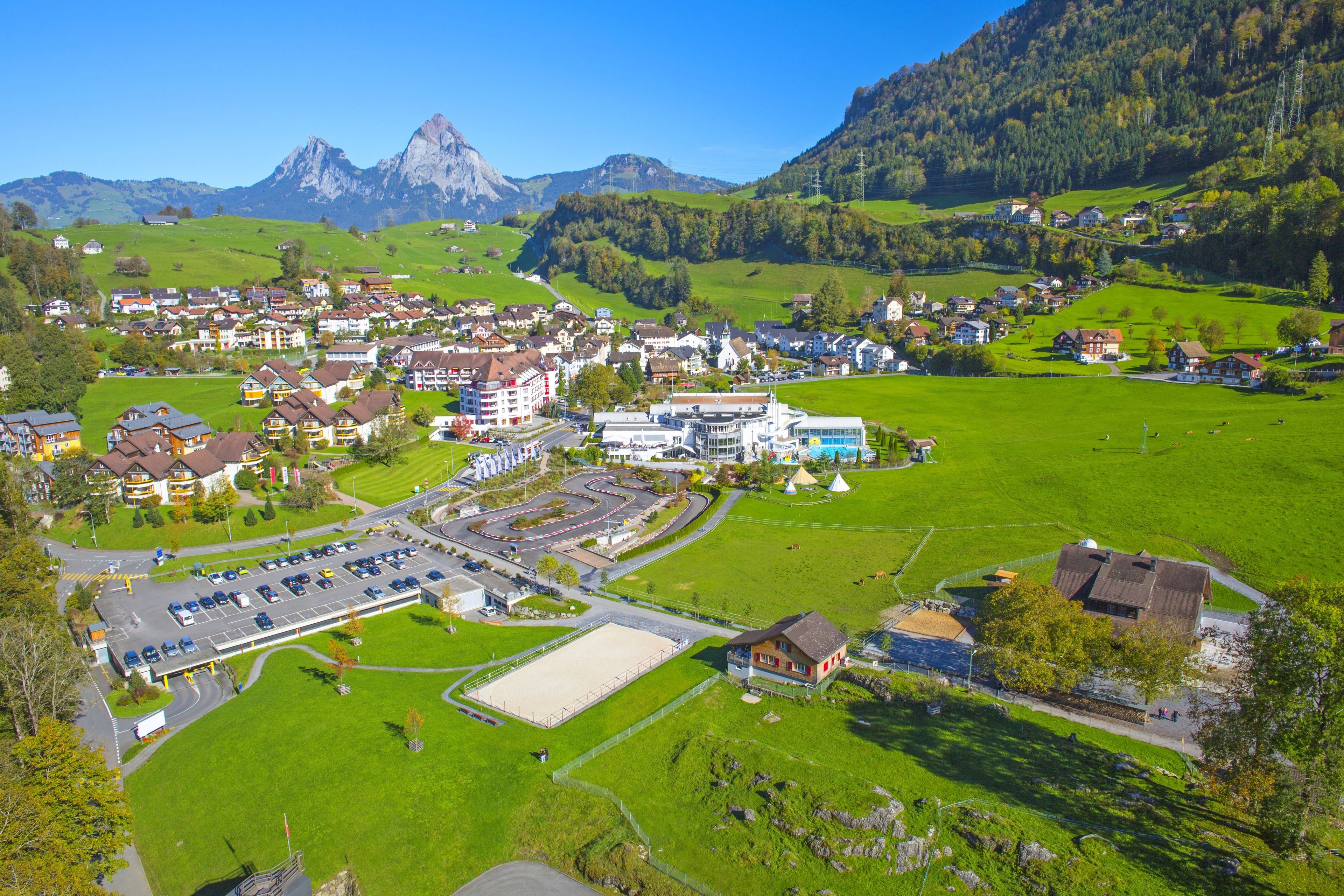 Eine Vogelperspektive zeigt eine Stadt mit einem zentralen Gebäudekomplex, umliegenden Häusern und einer Bergkulisse.