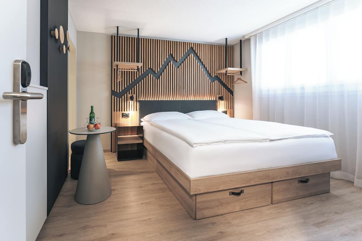 Ein modernes Hotelzimmer mit einem Bettgestell aus Holz, einem runden Tisch mit Flasche und Gläsern, einer Wandleuchte und einem großen Fenster mit transparenten Vorhängen.