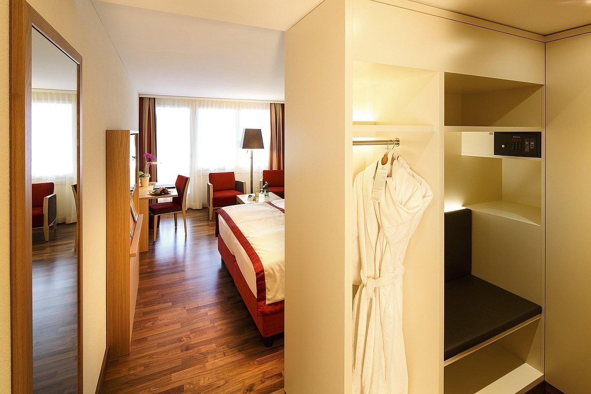Een hotelkamer met een houten vloer, een bed met rode beddengoed, een bureau met een stoel, een badjas hangend aan een rek, en een televisie op een plank.