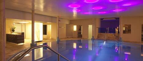 An indoor swimming pool with a staircase leading to a door, surrounded by walls and a ceiling with circular lights.