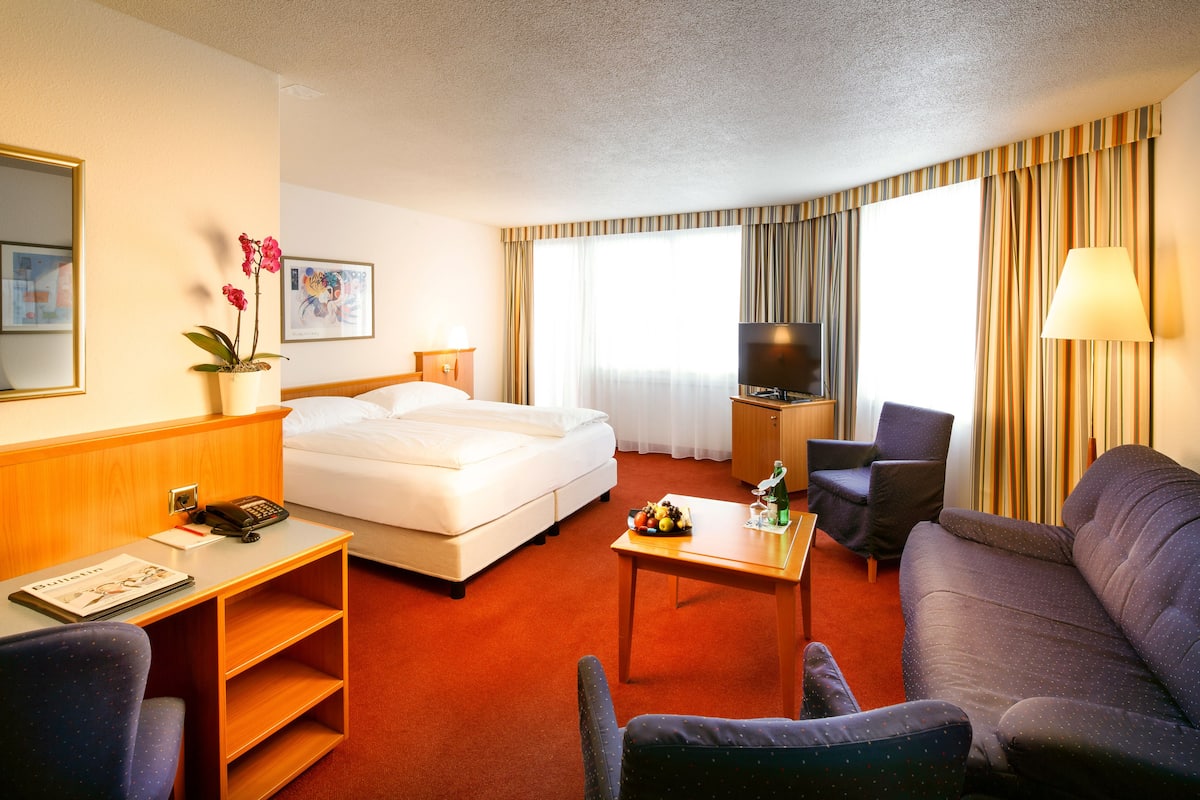 Ein Hotelzimmer mit Bett, einer Couch, einem Schreibtisch mit Lampe, einem Fernseher und einer Zimmerpflanze.