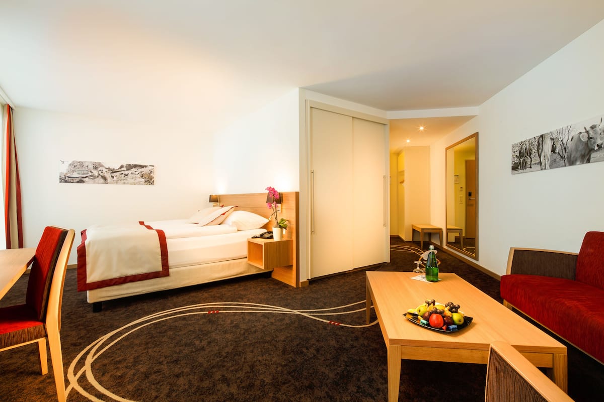 Ein Hotelzimmer mit einem Bett, einem Schreibtisch, einem Esstisch mit Obst, einer roten Couch und einem Spiegel.