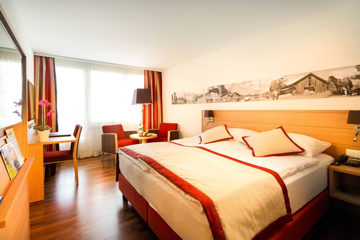 Ein Hotelzimmer mit einem großen Bett, einem Schreibtisch mit Stuhl, einem kleinen Tisch mit Lampe und einem Bilderrahmen auf dem Schreibtisch.