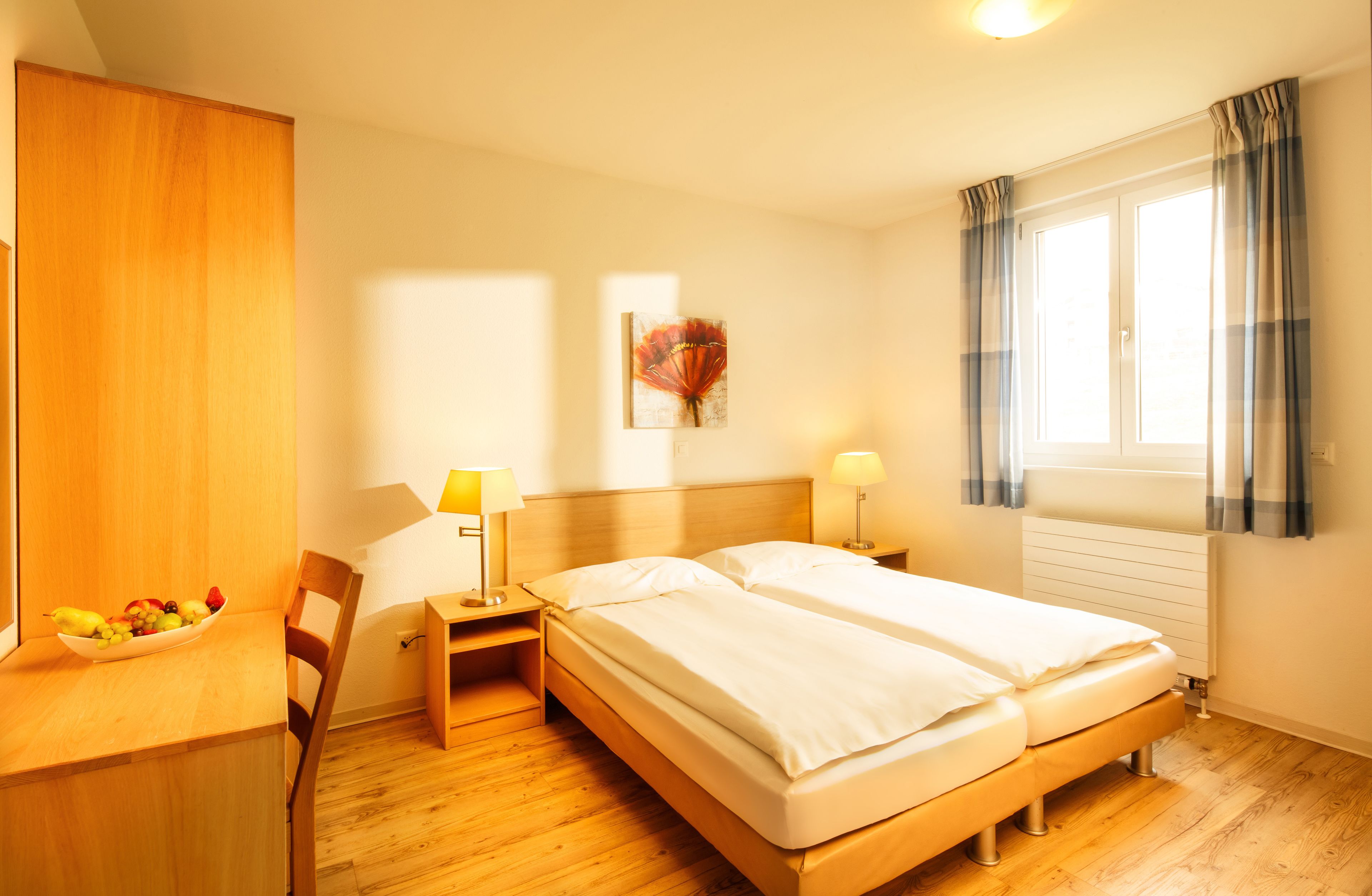 Ett hotellrum med två sängar, ett skrivbord i trä med en fruktskål, en stol, sänglampor, ett fönster med gardiner och ett konstverk som hänger på väggen.