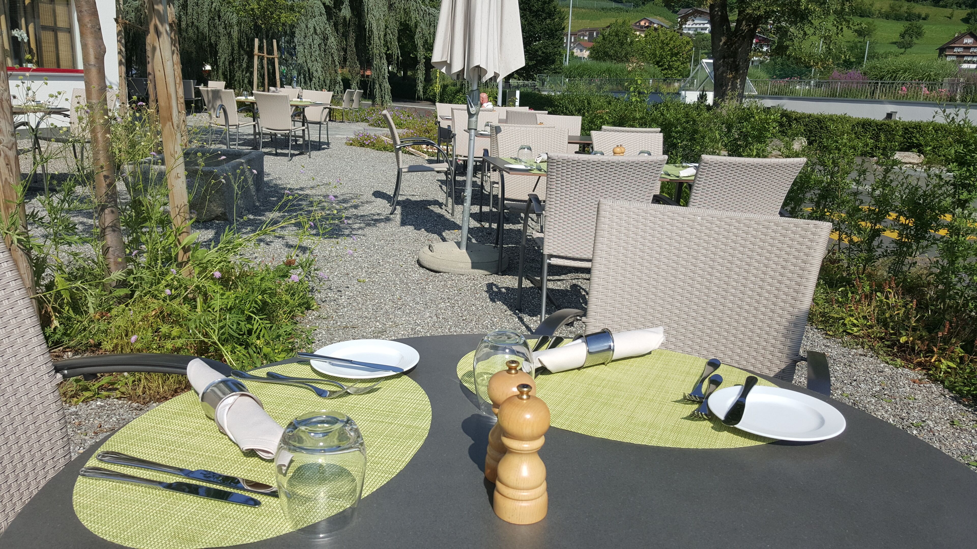 Un espace de restauration en plein air, avec des chaises et des tables en osier, entouré de verdure et offrant une vue sur une colline.