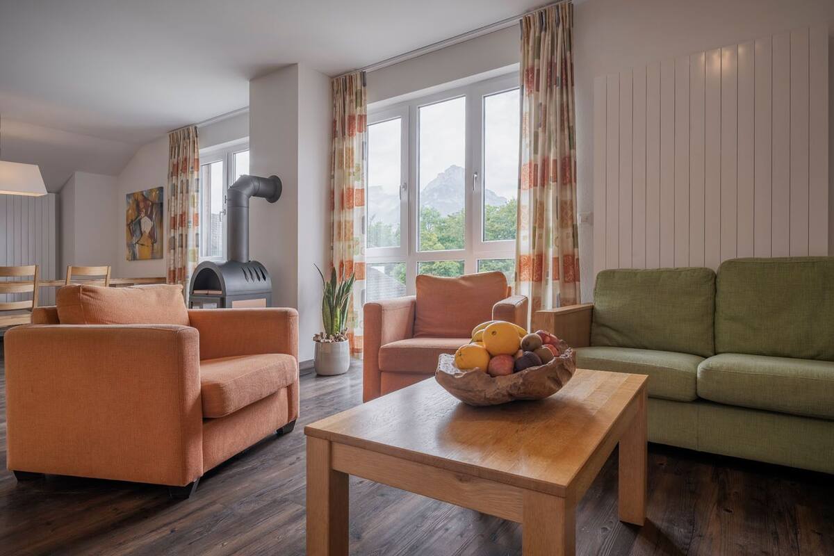 Ein modernes Wohnzimmer mit Kamin, orangefarbenen und grünen Sofas, einem Holztisch und einem Fenster mit Vorhängen.