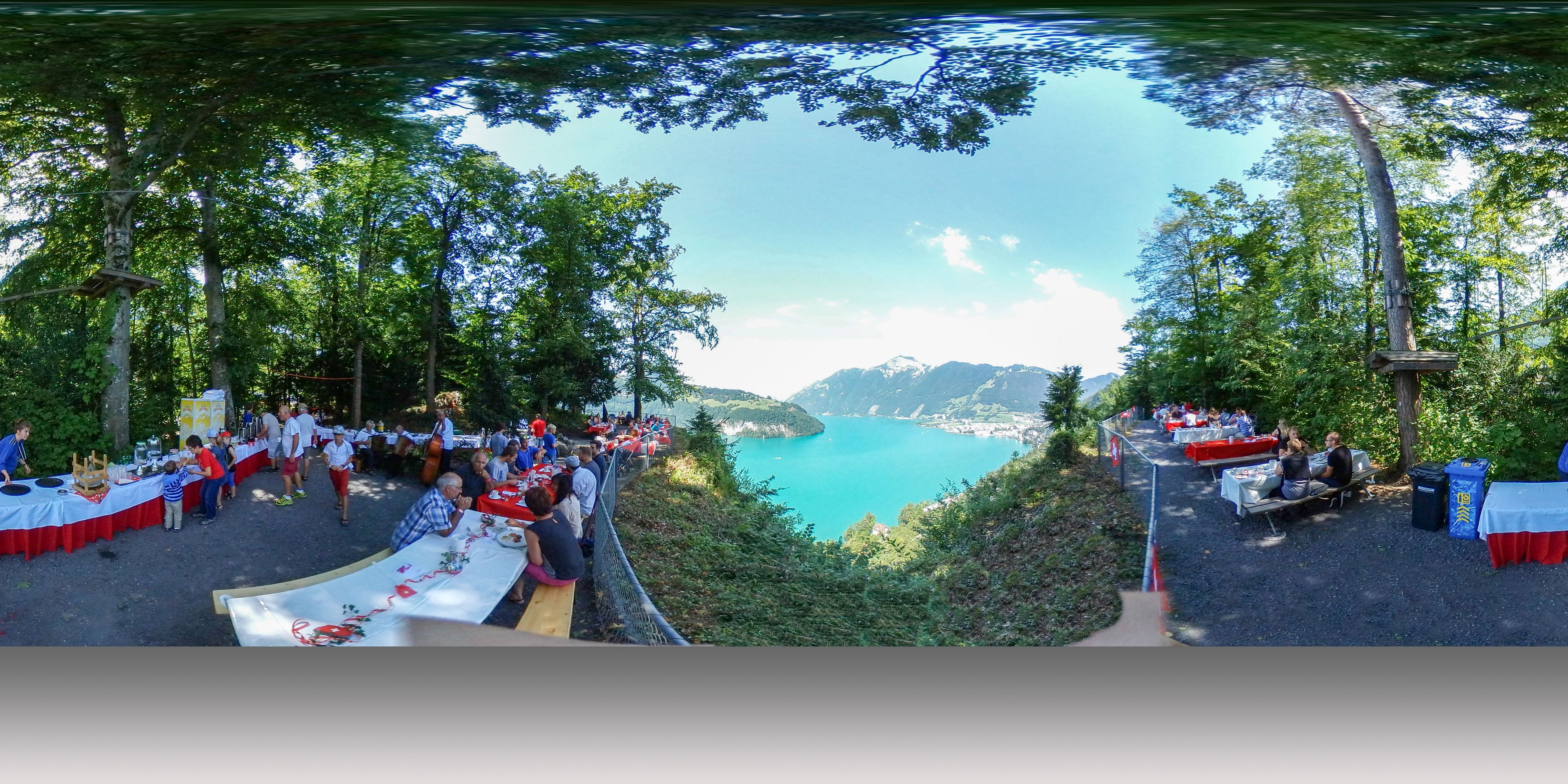 Vista panorámica de una reunión junto al lago, con mesas y sillas dispuestas para un evento, rodeado de árboles y con vistas a un lago tranquilo y montañas a lo lejos.