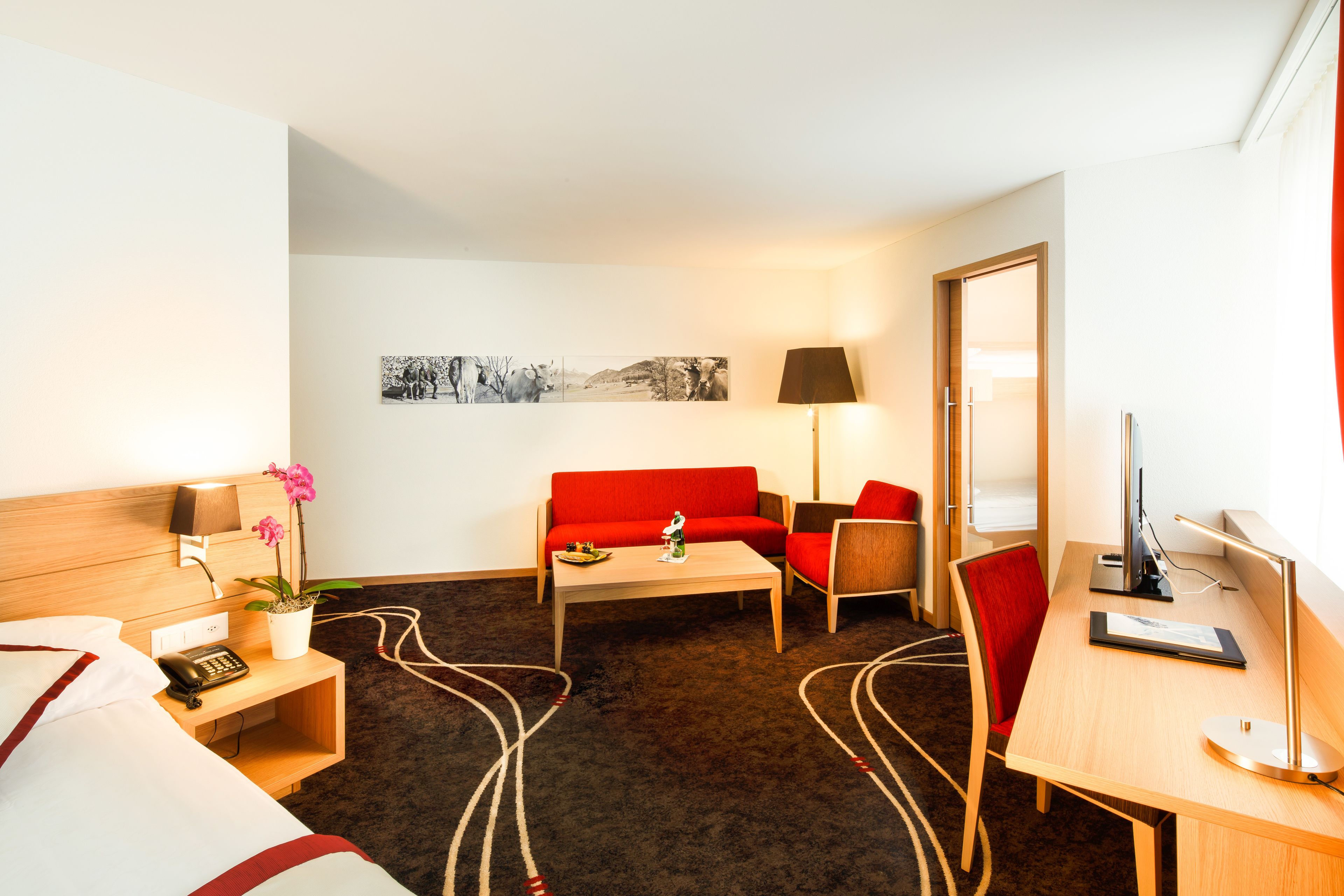 A hotel room with a bed, a red sofa, a wooden desk, a computer, a lamp, and a wall-mounted picture.