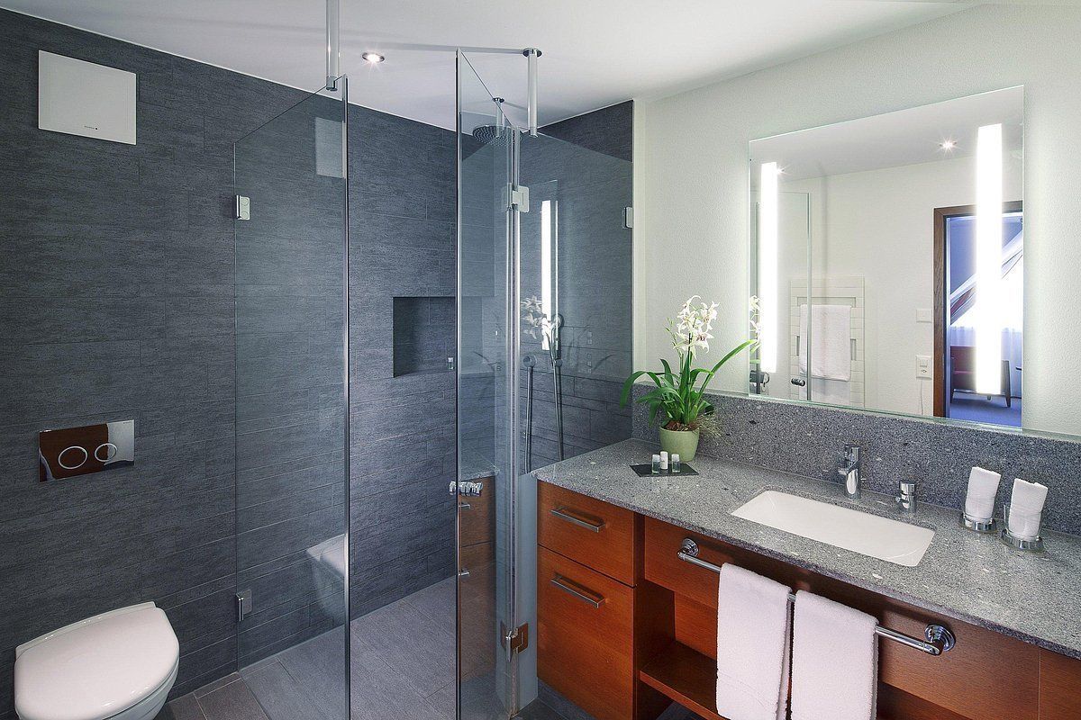 Un baño moderno con ducha envuelta en vidrio, inodoro blanco, lavamanos con encimera de granito y un espejo grande.