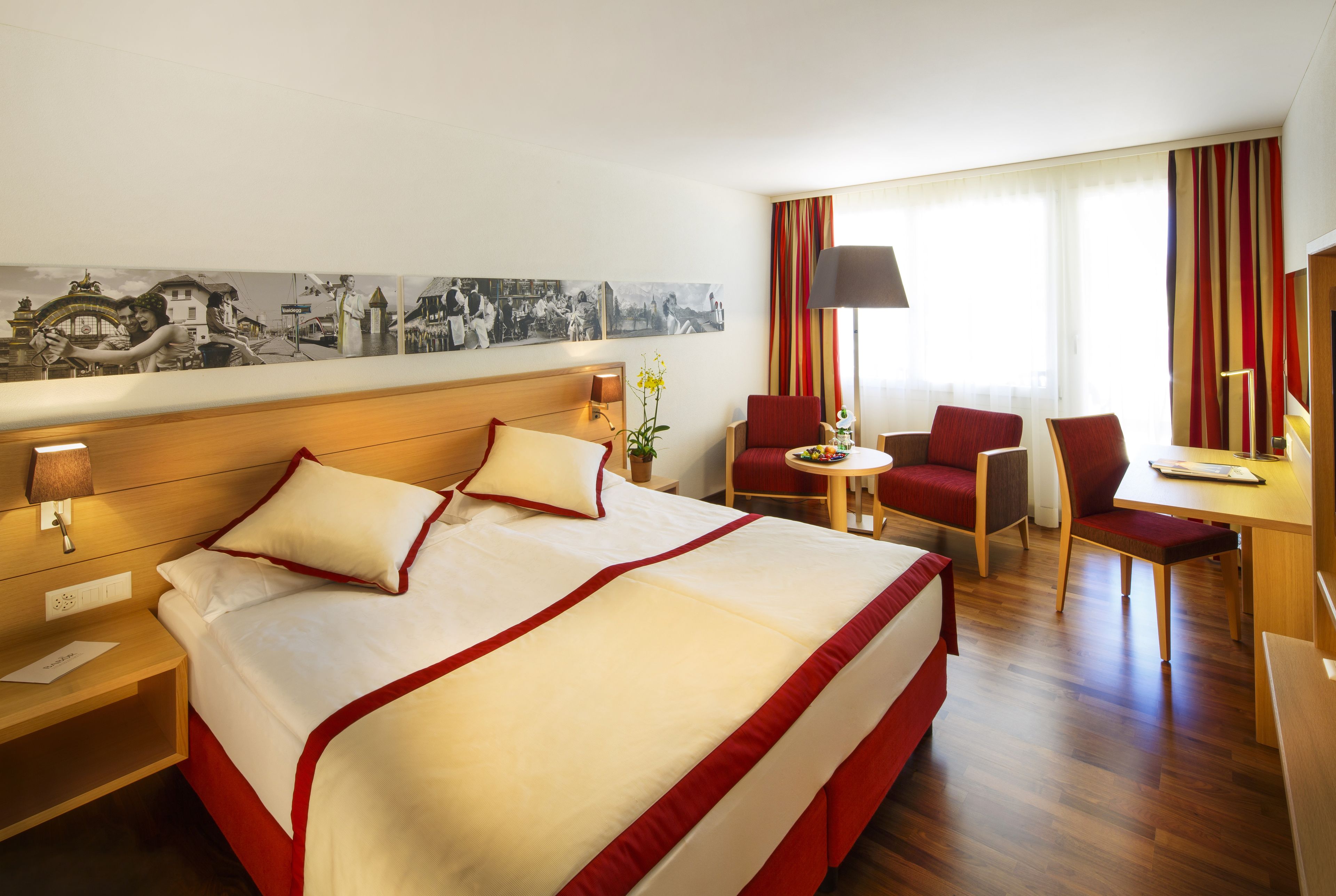 Habitación de hotel con una cama grande, un escritorio, dos sillas, una lámpara y un televisor montado en la pared.