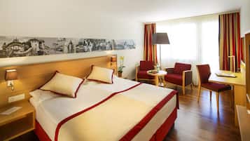 Habitación de hotel con una cama grande, un escritorio, dos sillas, una lámpara y un televisor montado en la pared.