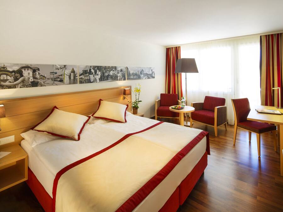 Habitación de hotel con una cama grande, un escritorio, dos sillas, una lámpara y un televisor montado en la pared.
