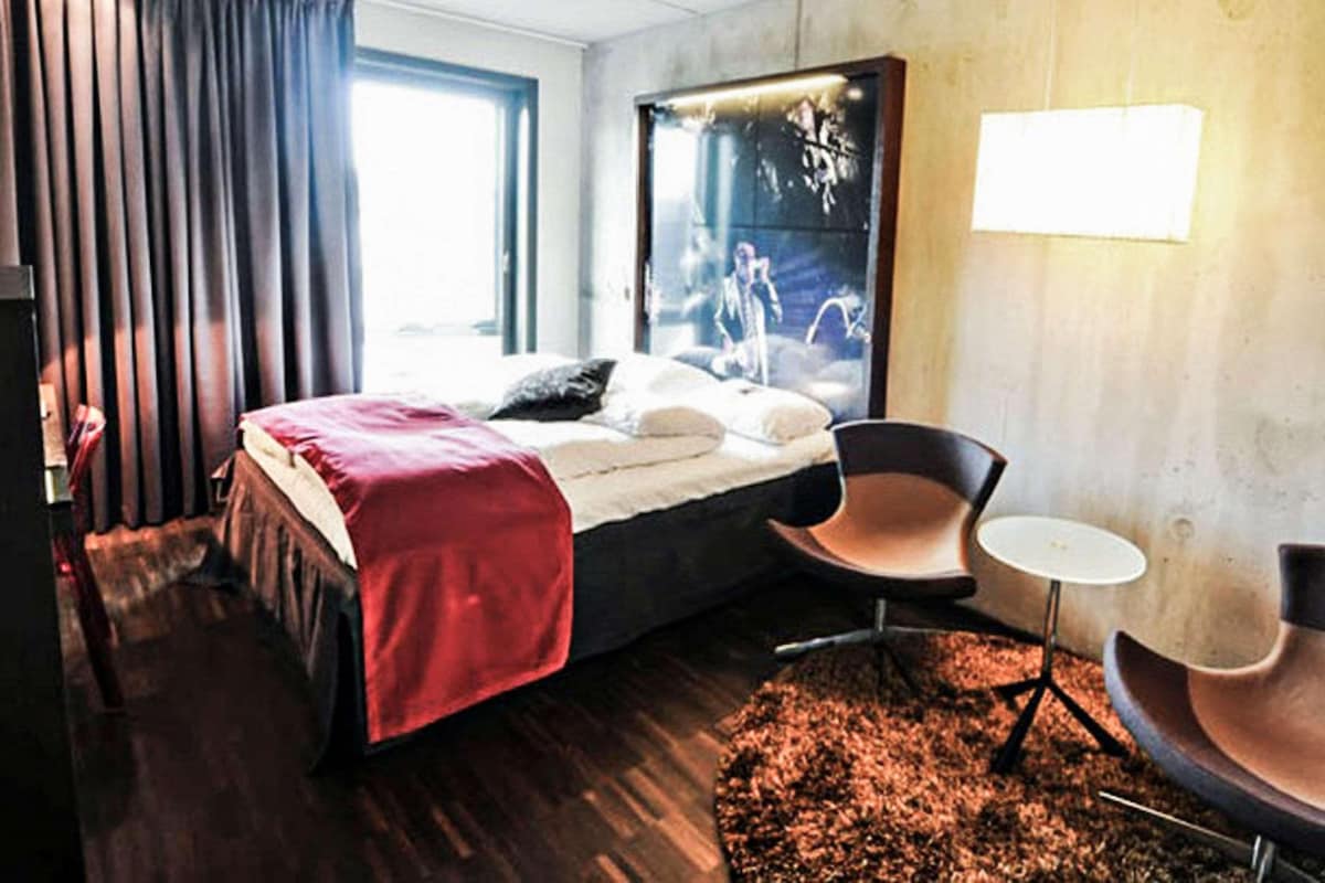 Una habitación de hotel moderna con una cama grande, dos sillas, una mesita y un ventanal amplio.