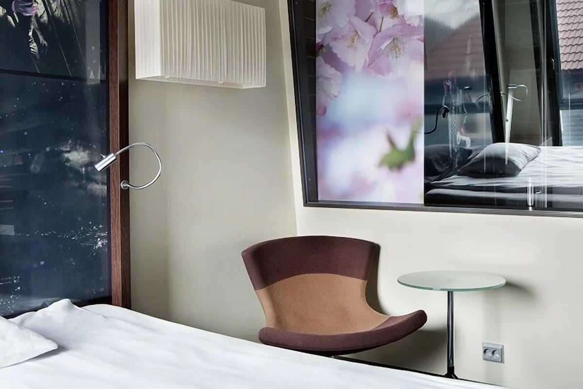 Habitación de hotel con cama, una silla, una mesita, ventana con vista a un edificio y una lámpara de pared.