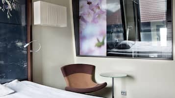 Une chambre d’hôtel avec un lit, une chaise, une petite table, une fenêtre donnant sur des fleurs et un climatiseur fixé au mur.