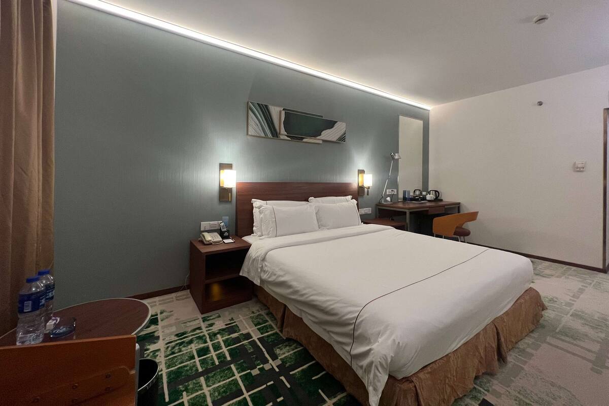 A hotel room with a bed, bedside tables, a desk, a chair, a mirror, and a wall-mounted artwork.