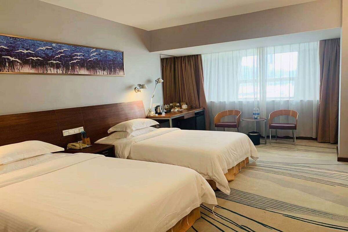 A hotel room with two beds, a desk with a lamp, a chair, a small table, and a large window with curtains.
