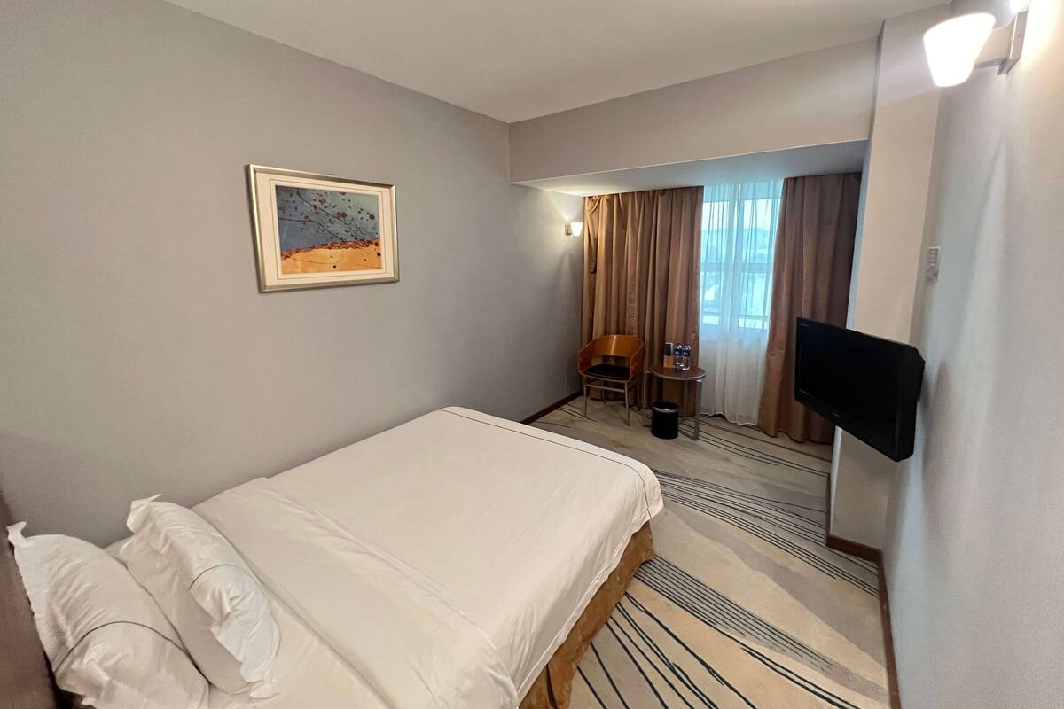 A hotel room with a bed, a chair, a small table with a bottle and glass, a television mounted on the wall, and a window with curtains.