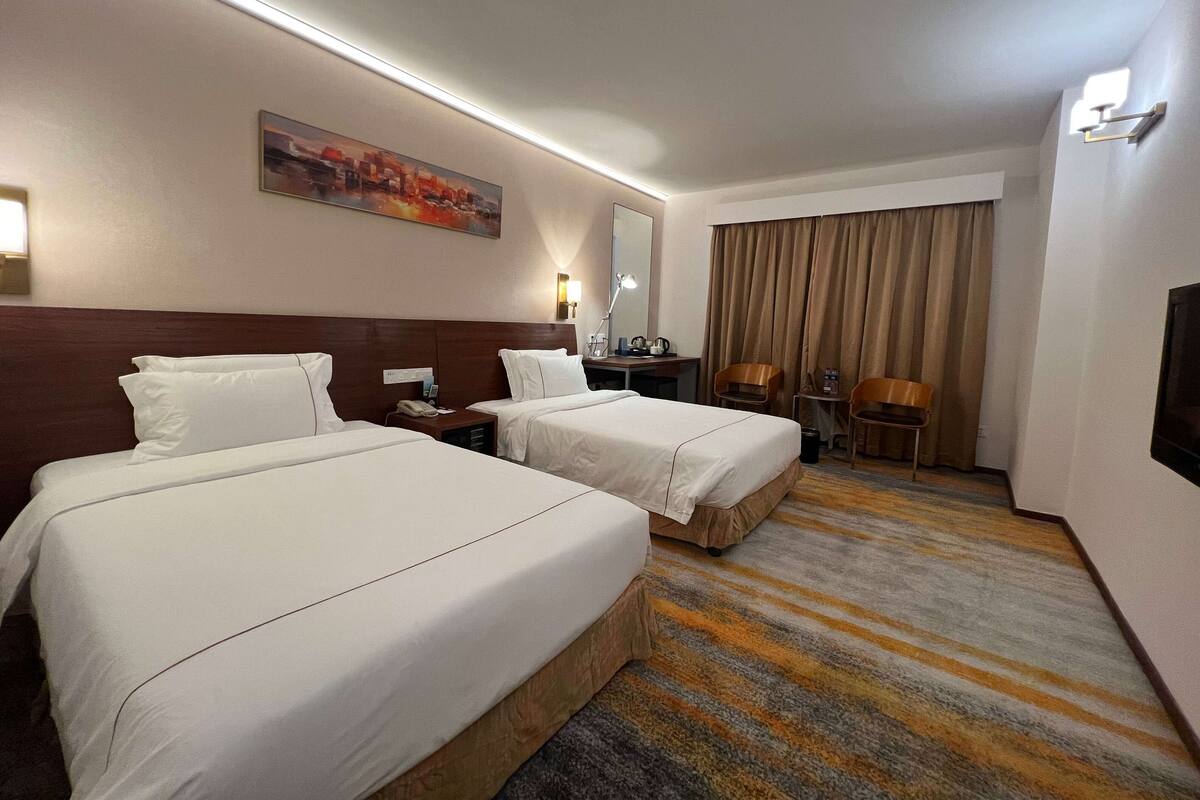 A hotel room with two beds, a desk with a chair, a lamp, and a large window with curtains.
