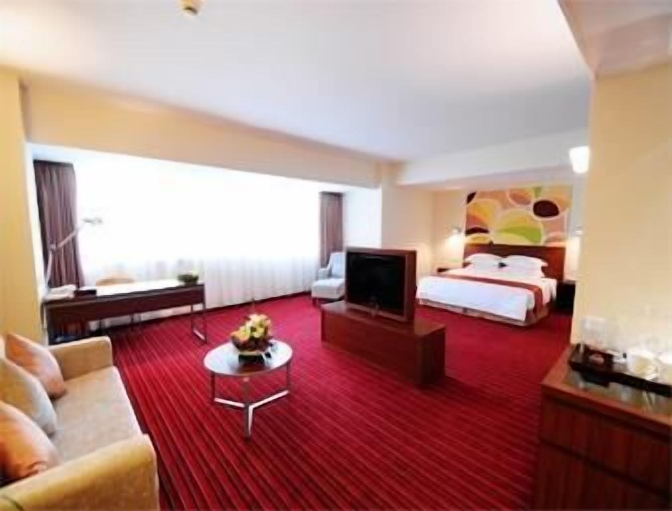 A hotel room with a red carpet, a bed, a TV, a desk, and a sofa.