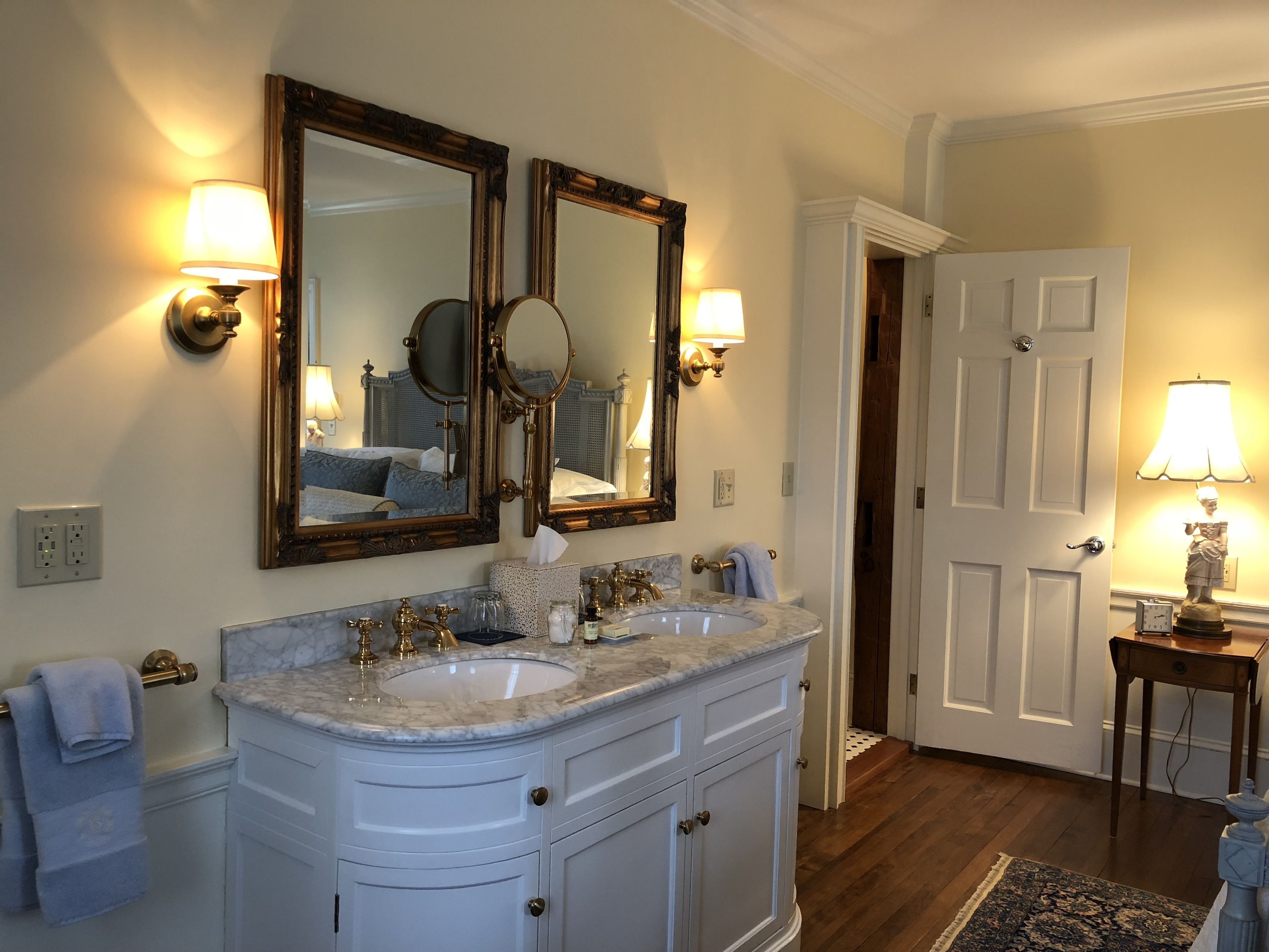 A bathroom with a double sink vanity, two wall-mounted mirrors, and a small table with a lamp.