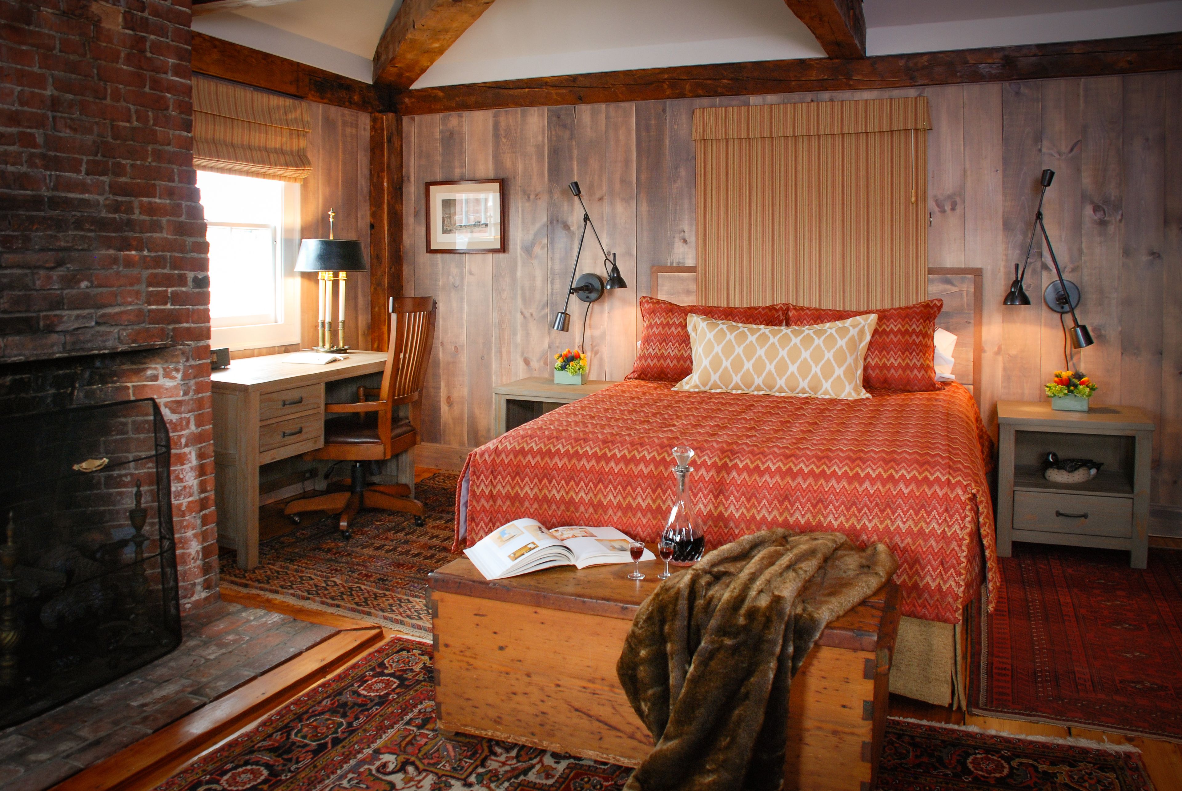 A cozy bedroom with a fireplace, a bed with patterned bedding, a desk with a lamp, and a nightstand with a lamp.