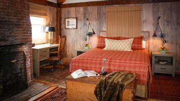 A cozy bedroom with a fireplace, a bed with patterned bedding, a desk with a lamp, and a nightstand with a lamp.