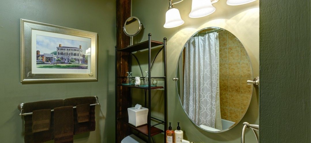 A bathroom with a framed painting, a towel rack, a shelf with toiletries, a round mirror, and a shower curtain.